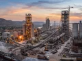 An elevated photograph of a large, complex cement plant operating at dusk. Flaring flames are visible from two of its rotary kilns, which are surrounded by towering structures and silos. Cranes and trucks are positioned around construction and production areas.