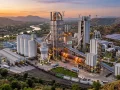 An aerial dusk photograph of a large, modern industrial plant with a rotary kiln and multiple tall buildings, located in a valley with a river and hills in India. The image highlights sustainable elements like solar panels and wind turbines, and includes a railway and truck depot